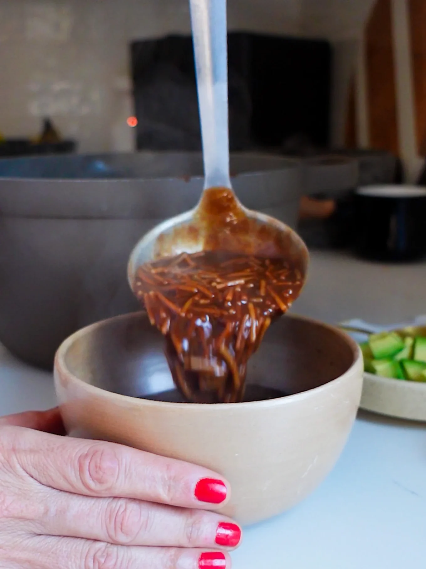 A hand with red nail polish holds a beige bowl while a ladle pours hot, dark brown Mexican soup into it. Steam rises from the soup. In the background, there is a bowl and a plate of chopped green vegetables on a kitchen counter.