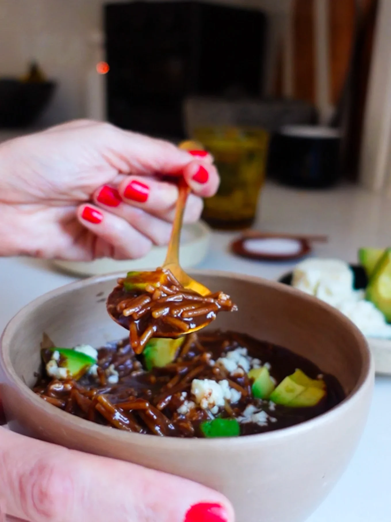 A close-up of a hand with red painted nails holding a spoonful of dark saucy noodles from a beige bowl, which also contains white rice and green avocado chunks—reminiscent of the cozy flavors found in classic Mexican soup.