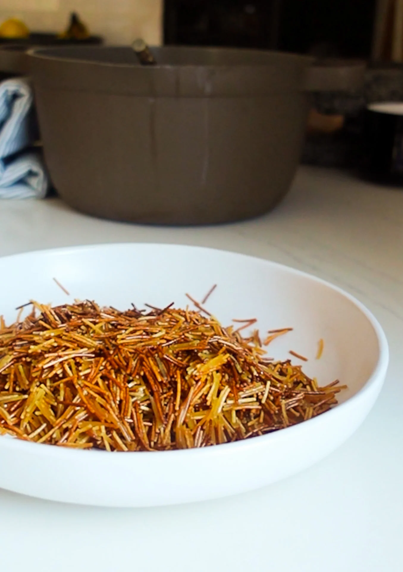 A white bowl filled with toasted brown and golden vermicelli noodles, perfect for Sopa de Fideo, sits on a white countertop. In the blurred background, there is a large gray pot, a blue-striped towel, and some kitchen items.