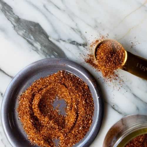A small blue-gray plate with a swirl of reddish-brown homemade spice blend sits on a white marble surface. Nearby, a gold measuring spoon filled with more of the seasoning and a glass jar, partially visible, complete the composition.