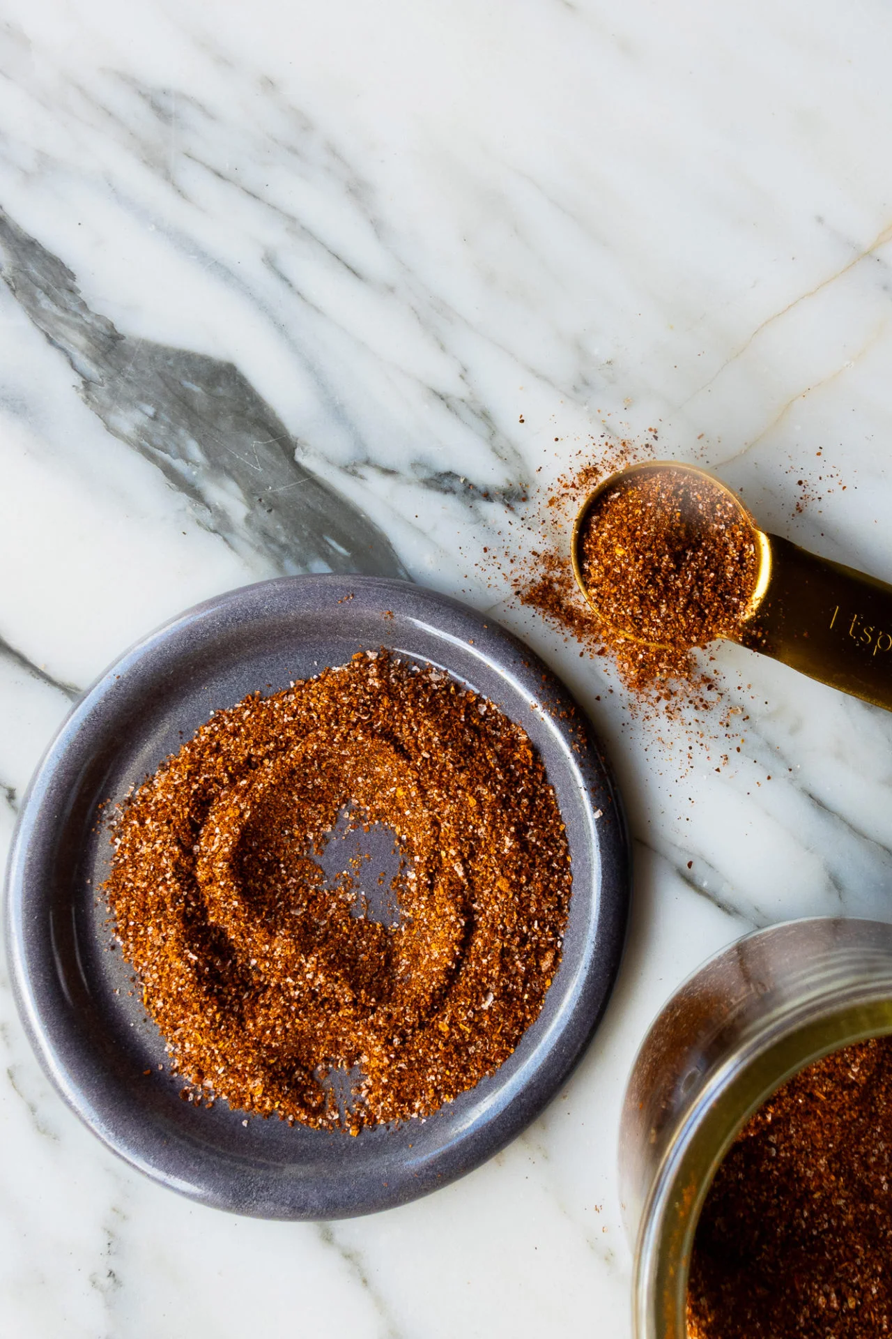 A small blue-gray plate with a swirl of reddish-brown homemade spice blend sits on a white marble surface. Nearby, a gold measuring spoon filled with more of the seasoning and a glass jar, partially visible, complete the composition.