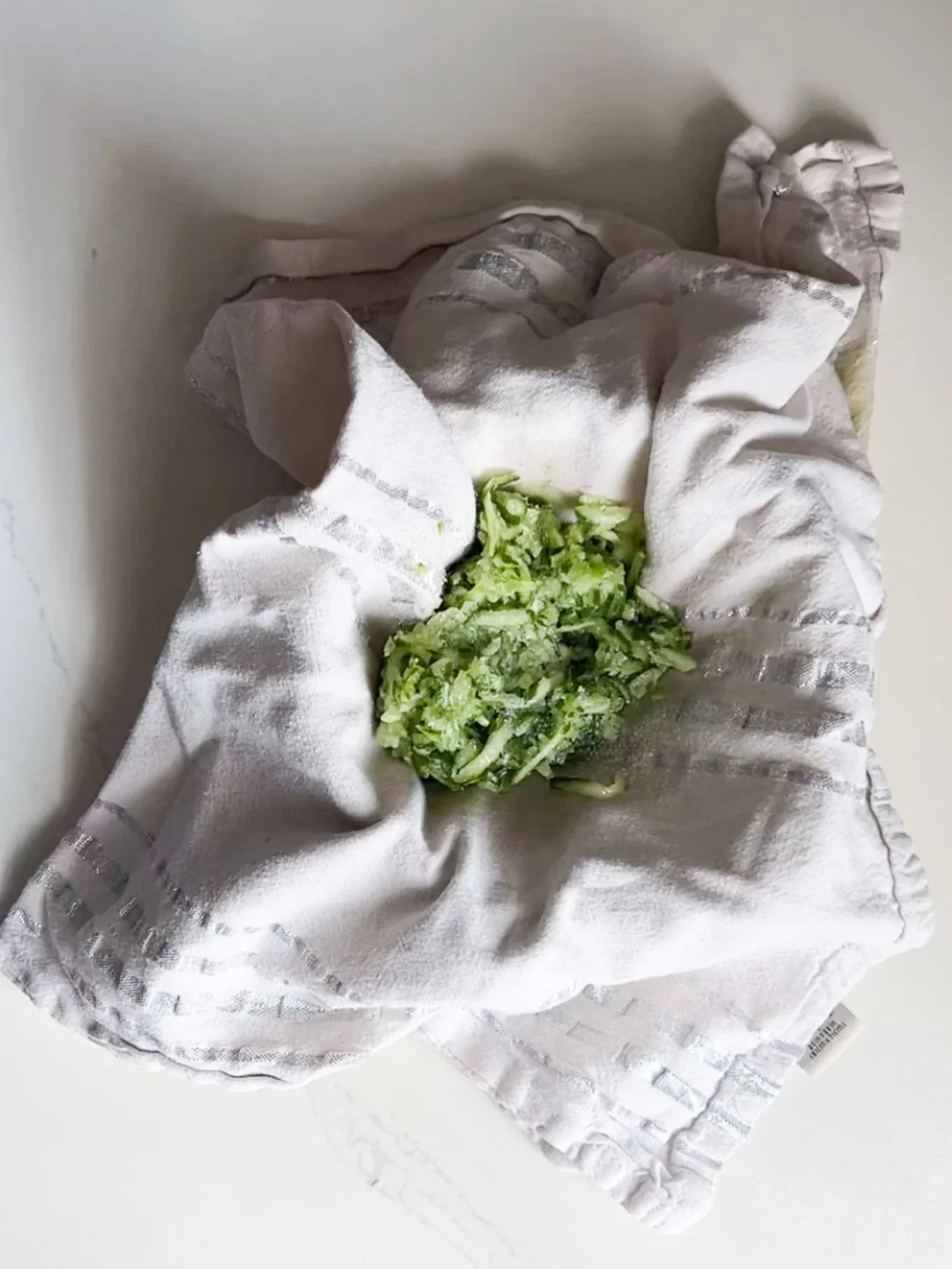 A mound of shredded zucchini sits on a light-colored kitchen towel with gray stripes, placed on a white countertop—perfect for preparing a fresh Tzatziki recipe or creamy Greek yogurt sauce. The towel is gathered loosely around the zucchini, forming a bowl-like shape.