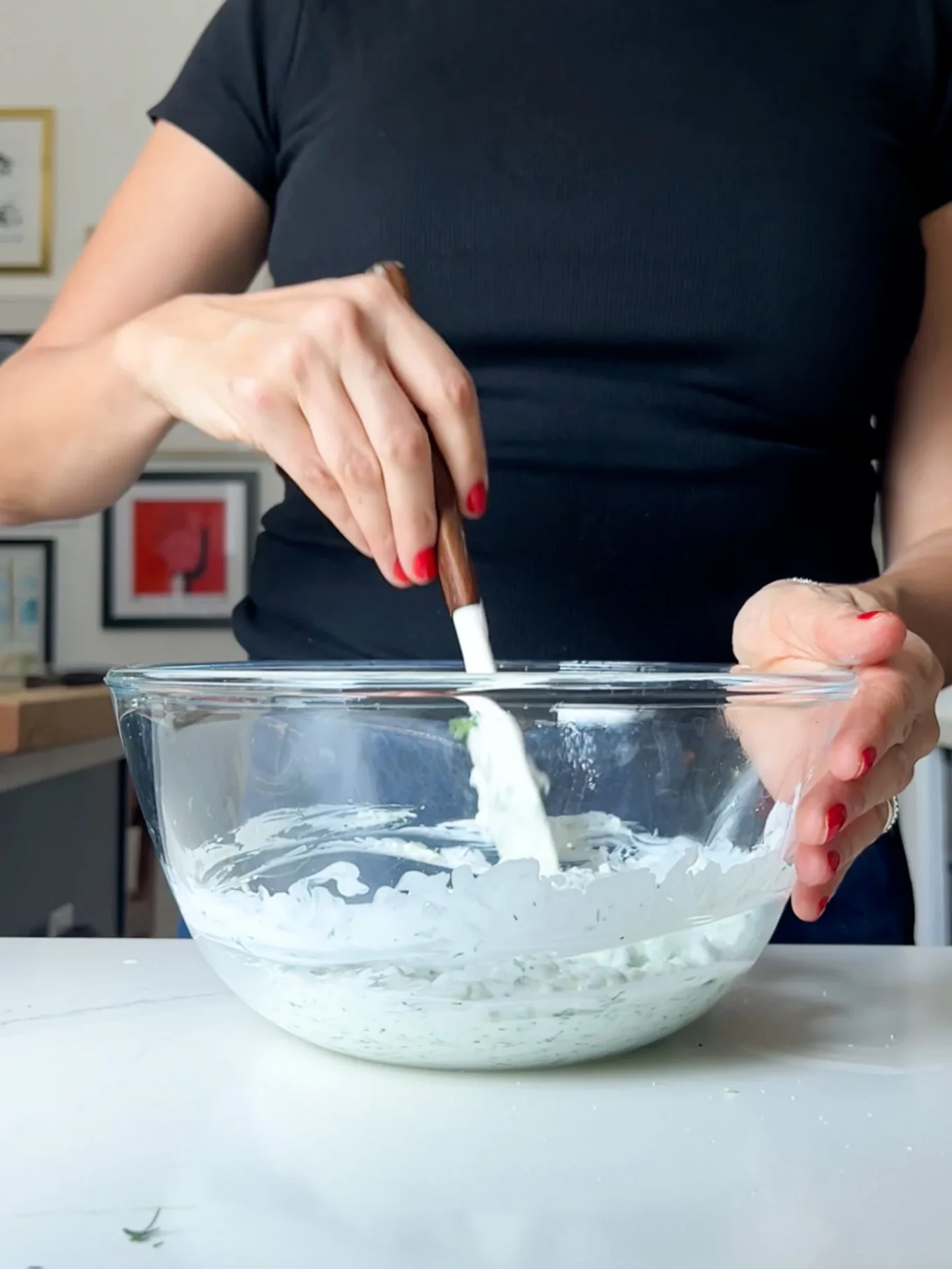 A person wearing a black short-sleeve shirt mixes a creamy Greek yogurt Tzatziki recipe in a large glass bowl using a spatula. The bowl sits on a white countertop, red nails visible, with framed art on the wall in the background.