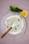A glass bowl with creamy Greek yogurt sauce, mixed with chopped dill, sits on a pink surface. A wooden-handled spoon rests inside. Nearby are four mini cucumbers and a halved lemon with visible seeds.