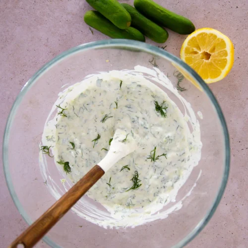 A glass bowl with creamy Greek yogurt sauce, mixed with chopped dill, sits on a pink surface. A wooden-handled spoon rests inside. Nearby are four mini cucumbers and a halved lemon with visible seeds.