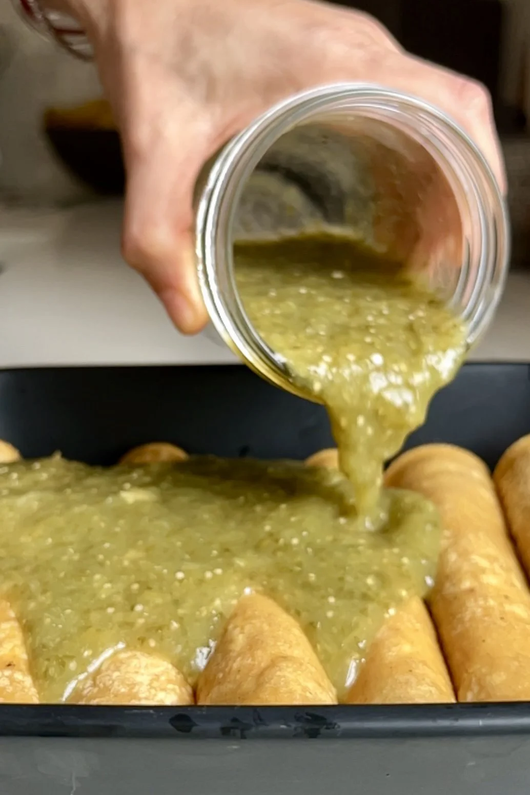 A hand pours thick green salsa verde from a glass jar over a row of pulled pork enchiladas in a black baking dish, coating the tops evenly. The enchiladas are arranged closely together and ready for baking on a kitchen countertop.