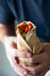 A person wearing a navy blue shirt holds a Vegetarian Gyro pita wrap filled with grilled vegetables and greens. The pita, partially wrapped in brown paper, includes red bell peppers, onions, lettuce, and tender King Oyster Mushrooms. The background is softly blurred.