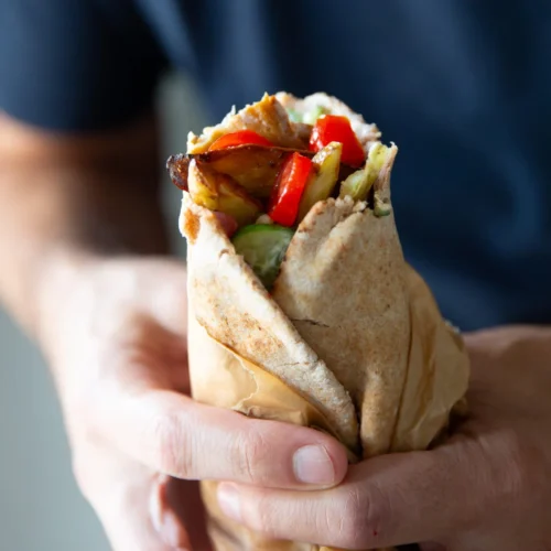 A person wearing a navy blue shirt holds a Vegetarian Gyro pita wrap filled with grilled vegetables and greens. The pita, partially wrapped in brown paper, includes red bell peppers, onions, lettuce, and tender King Oyster Mushrooms. The background is softly blurred.