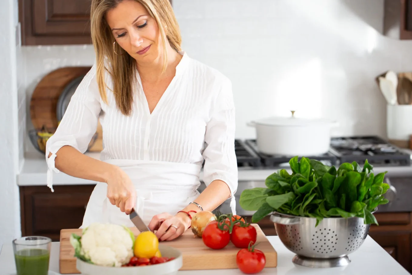 A woman in a white blouse chops an onion on a wooden cutting board in a bright kitchen. Fresh vegetables, including tomatoes, cauliflower, and spinach, are on the counter nearby. A green drink is beside her, and sunlight streams in from the left.
