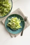 A teal plate with a serving of coleslaw made of shredded cabbage and lettuce sits on a beige napkin next to a silver fork, with a large glass bowl of Ranch Chopped Salad partially visible in the upper left corner on a white surface.