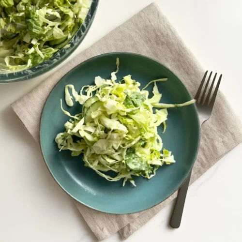 A teal plate with a serving of coleslaw made of shredded cabbage and lettuce sits on a beige napkin next to a silver fork, with a large glass bowl of Ranch Chopped Salad partially visible in the upper left corner on a white surface.