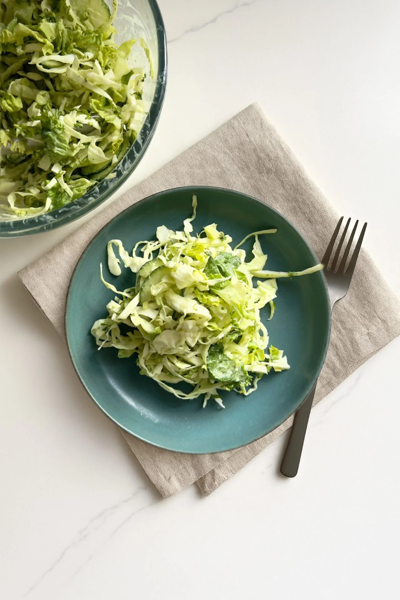 A teal plate with a serving of coleslaw made of shredded cabbage and lettuce sits on a beige napkin next to a silver fork, with a large glass bowl of Ranch Chopped Salad partially visible in the upper left corner on a white surface.