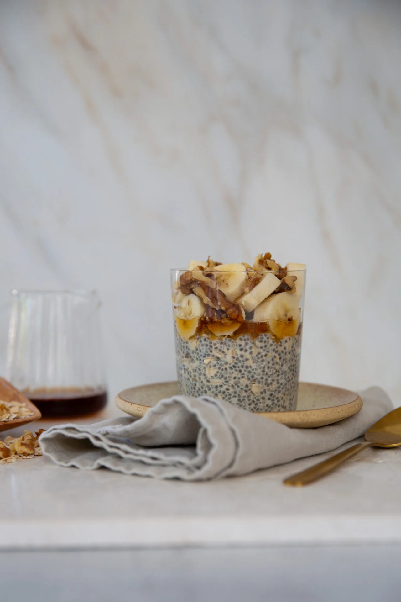 A glass filled with chia pudding topped with banana slices, chopped nuts, and a drizzle of syrup sits on a beige plate. A folded gray napkin and a gold spoon are in front, with a small glass jar of syrup in the background on a light marble surface.