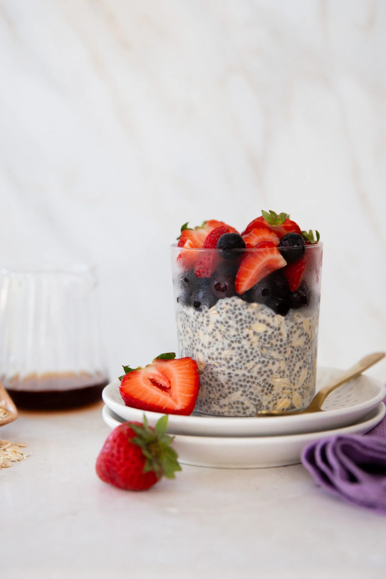 A glass filled with creamy overnight oats and chia seeds, topped with fresh berries like sliced strawberries and blueberries, sits on stacked white plates. A spoon rests nearby, alongside a vibrant strawberry and a purple napkin.