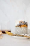 A clear glass filled with chia pudding, topped with yogurt, peanut butter, chocolate chips, and a drizzle of syrup sits on a saucer with a gold spoon. There’s a linen napkin, scattered oats, and a glass jar of dark syrup in the background.