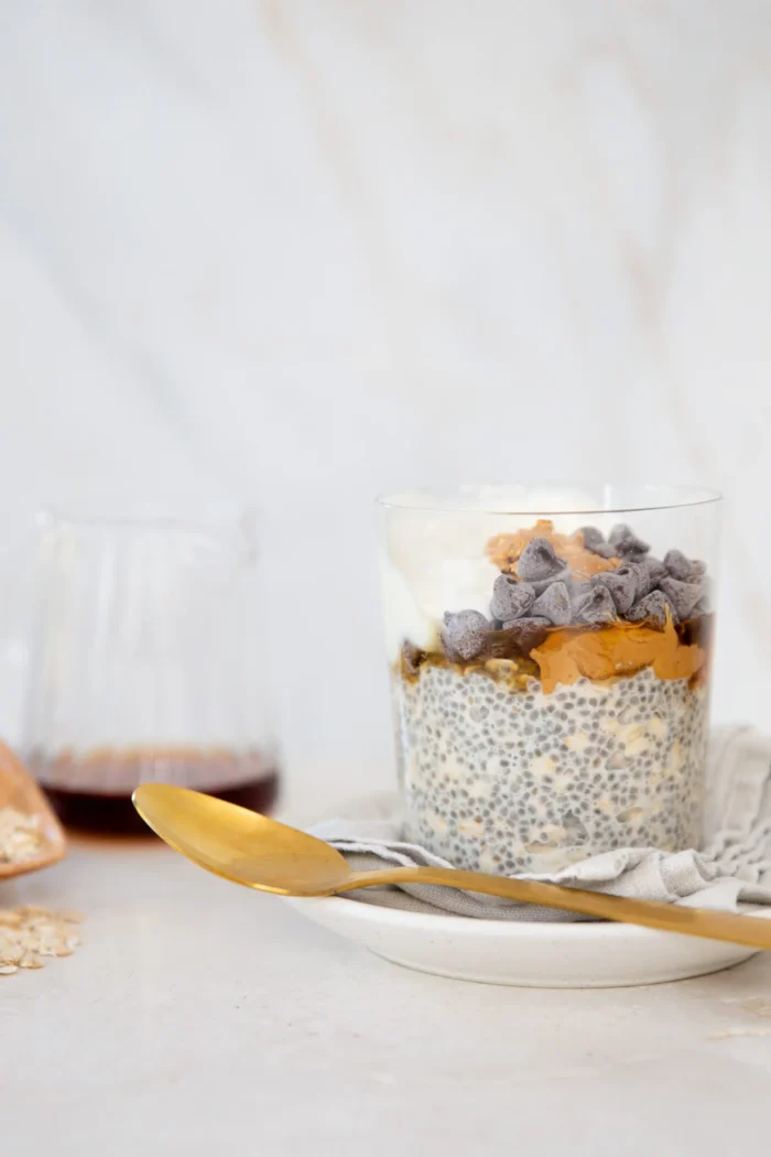 A clear glass filled with chia pudding, topped with yogurt, peanut butter, chocolate chips, and a drizzle of syrup sits on a saucer with a gold spoon. There’s a linen napkin, scattered oats, and a glass jar of dark syrup in the background.