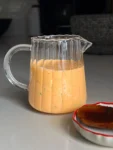 A clear glass pitcher filled with a creamy orange sauce sits on a light gray countertop; the sauce appears speckled with herbs. In the foreground, part of a small dish with a glossy brown sauce is visible. The background is softly out of focus.