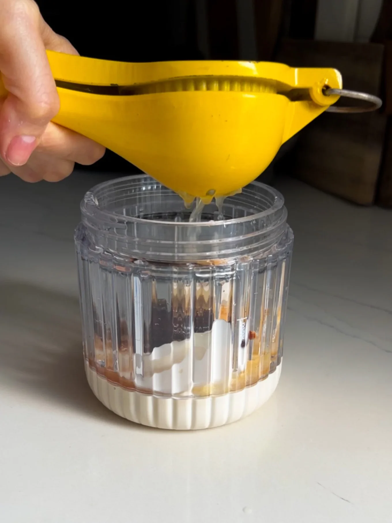 A hand uses a yellow citrus squeezer to press juice into a clear container filled with white liquid and brownish ingredients on a light countertop, suggesting an easy Chipotle Ranch Dressing homemade recipe in the making.