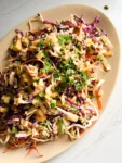 An oval plate holds a vibrant Chipotle Ranch Cabbage Salad with shredded green and purple cabbage, carrots, cucumber, cilantro, edamame, and crunchy pepitas, all tossed in a creamy dressing and served on a white marble surface.