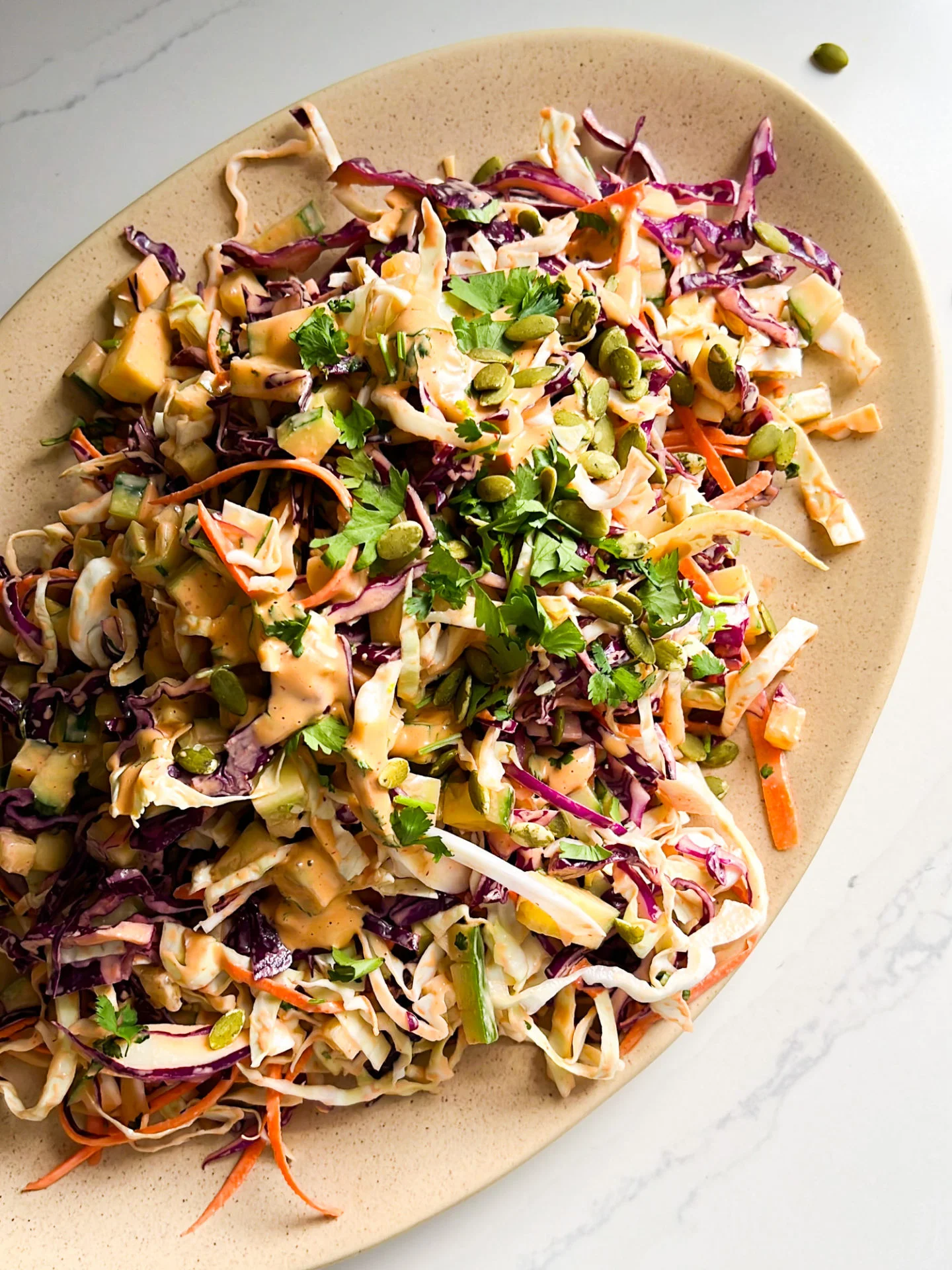An oval plate holds a vibrant Chipotle Ranch Cabbage Salad with shredded green and purple cabbage, carrots, cucumber, cilantro, edamame, and crunchy pepitas, all tossed in a creamy dressing and served on a white marble surface.