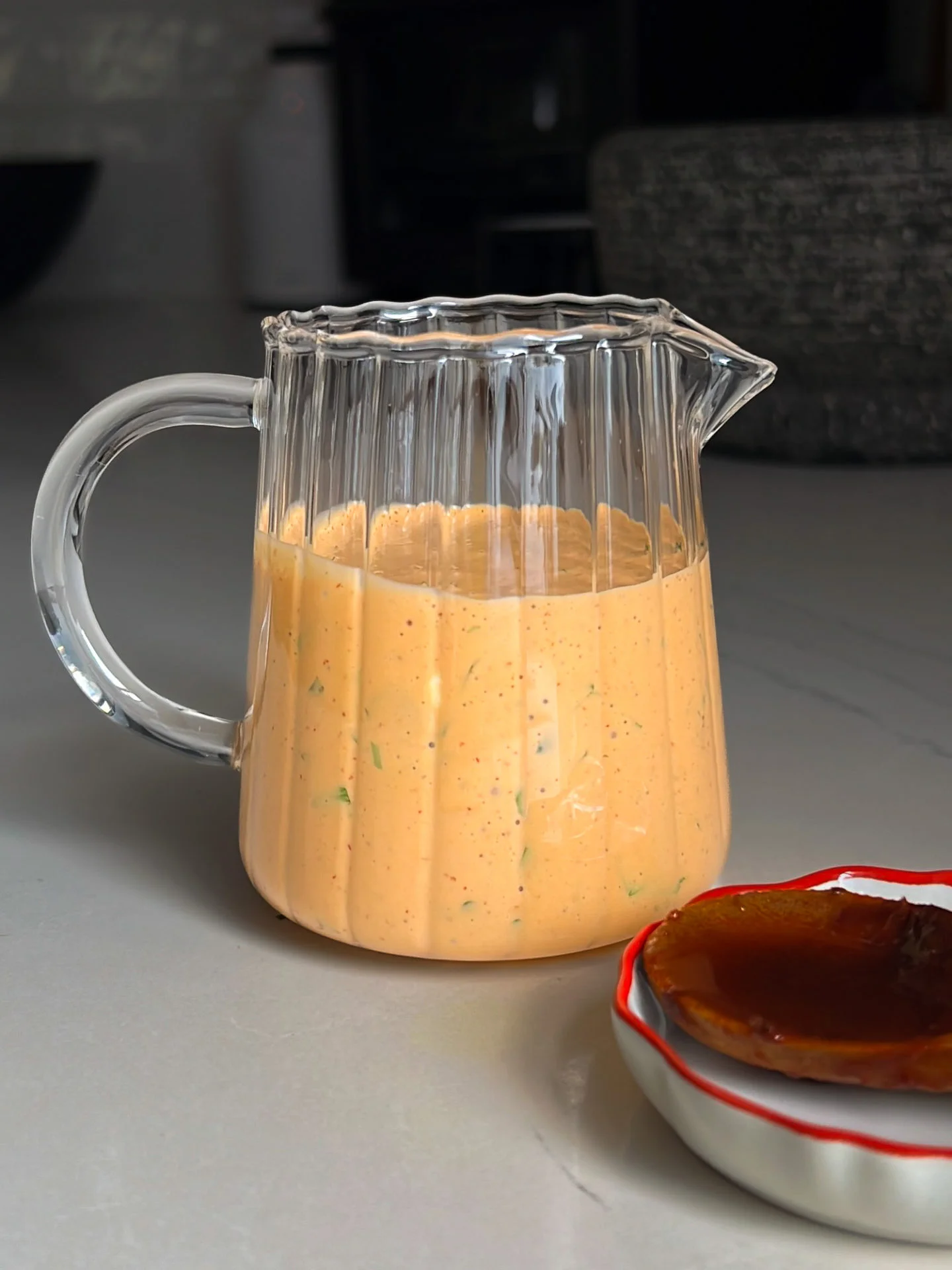 A clear glass pitcher filled with a creamy orange sauce sits on a light gray countertop; the sauce appears speckled with herbs. In the foreground, part of a small dish with a glossy brown sauce is visible. The background is softly out of focus.
