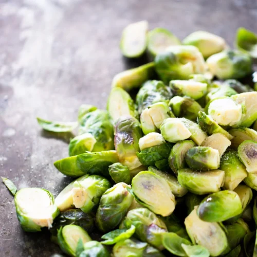 A pile of fresh Brussels sprouts, perfect for vegan brussel sprout recipes, rests on a dark, textured surface. Their vibrant green hues and natural leafy details catch the light along their cut edges.