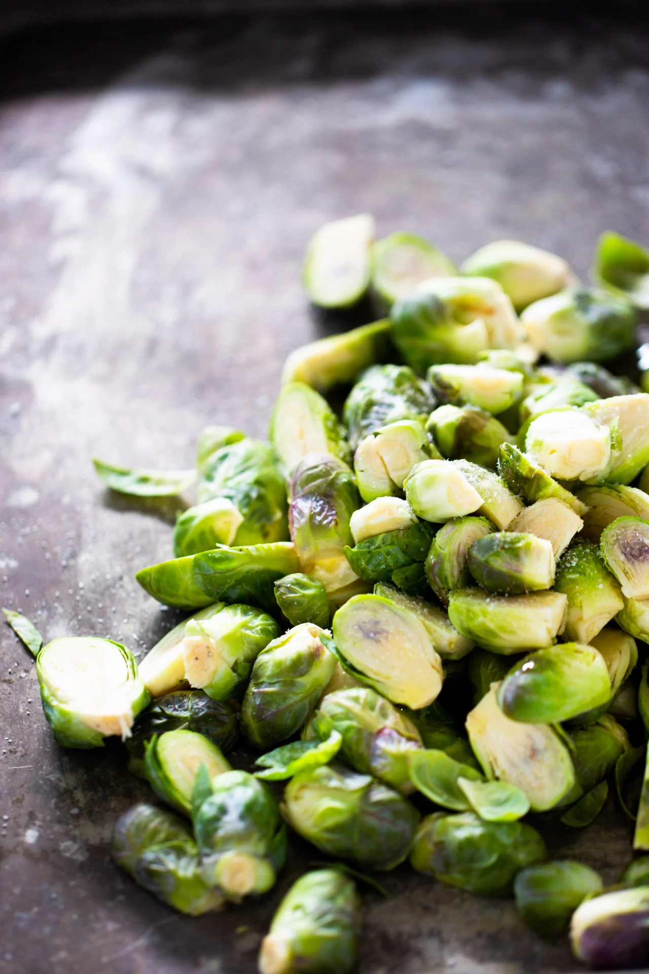 A pile of fresh Brussels sprouts, perfect for vegan brussel sprout recipes, rests on a dark, textured surface. Their vibrant green hues and natural leafy details catch the light along their cut edges.