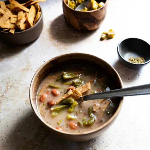 A brown bowl of Sopa de Frijol with visible okra and tomatoes sits on a countertop. Two spoons rest in the bowl. Nearby are crispy tortilla strips, chopped green pickles, and dried herbs—all bathed in warm, natural lighting.