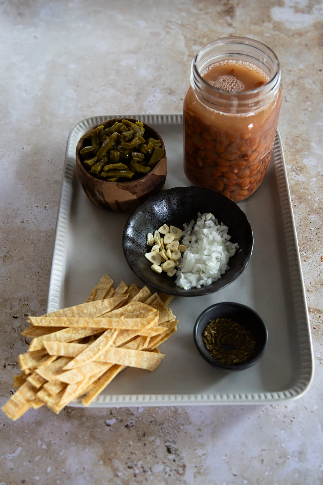 A tray holds julienned tortilla strips, sliced jalapeños in a bowl, chopped onion, sliced garlic, jarred pinto beans in broth for Sopa de Frijol, and a small bowl of dried oregano—perfect for a receta fácil on a light-colored surface.