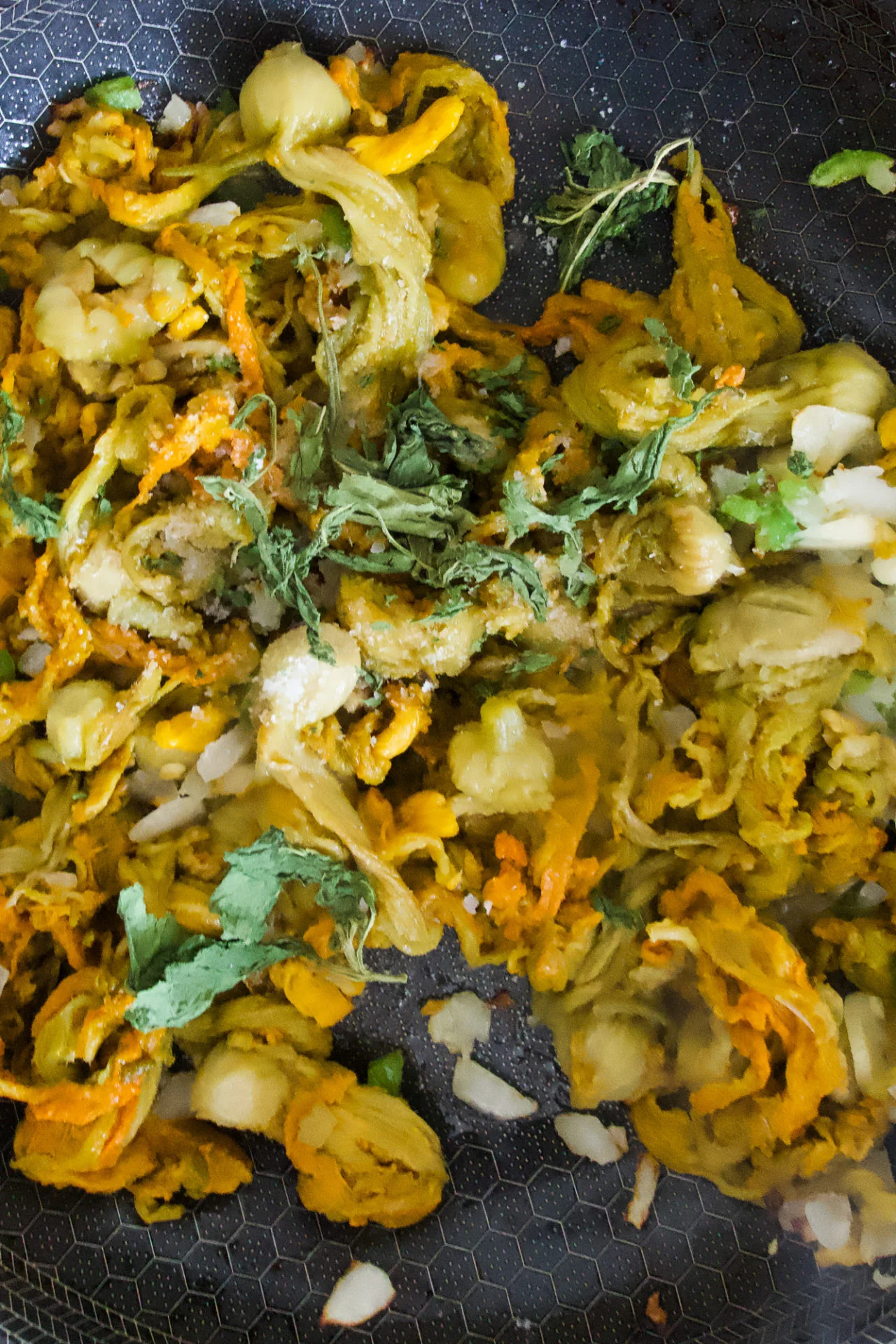 Close-up of cooked yellow and green vegetables, likely squash blossom and chopped greens, garnished with fresh cilantro. The mixture, perfect for Squash Blossom Quesadillas (Quesadillas de Flor de Calabaza), sits in a non-stick pan with a hexagonal pattern.