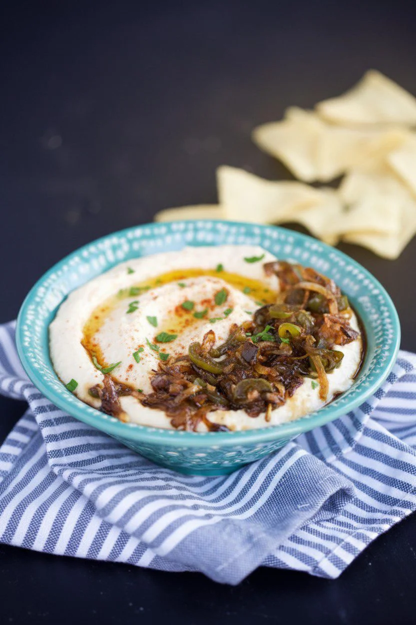 A turquoise bowl filled with creamy Mexican hummus, garnished with sautéed onions and green peppers, sits on a folded blue-striped cloth. The bowl rests on a dark surface, with pieces of pita bread in the background.
