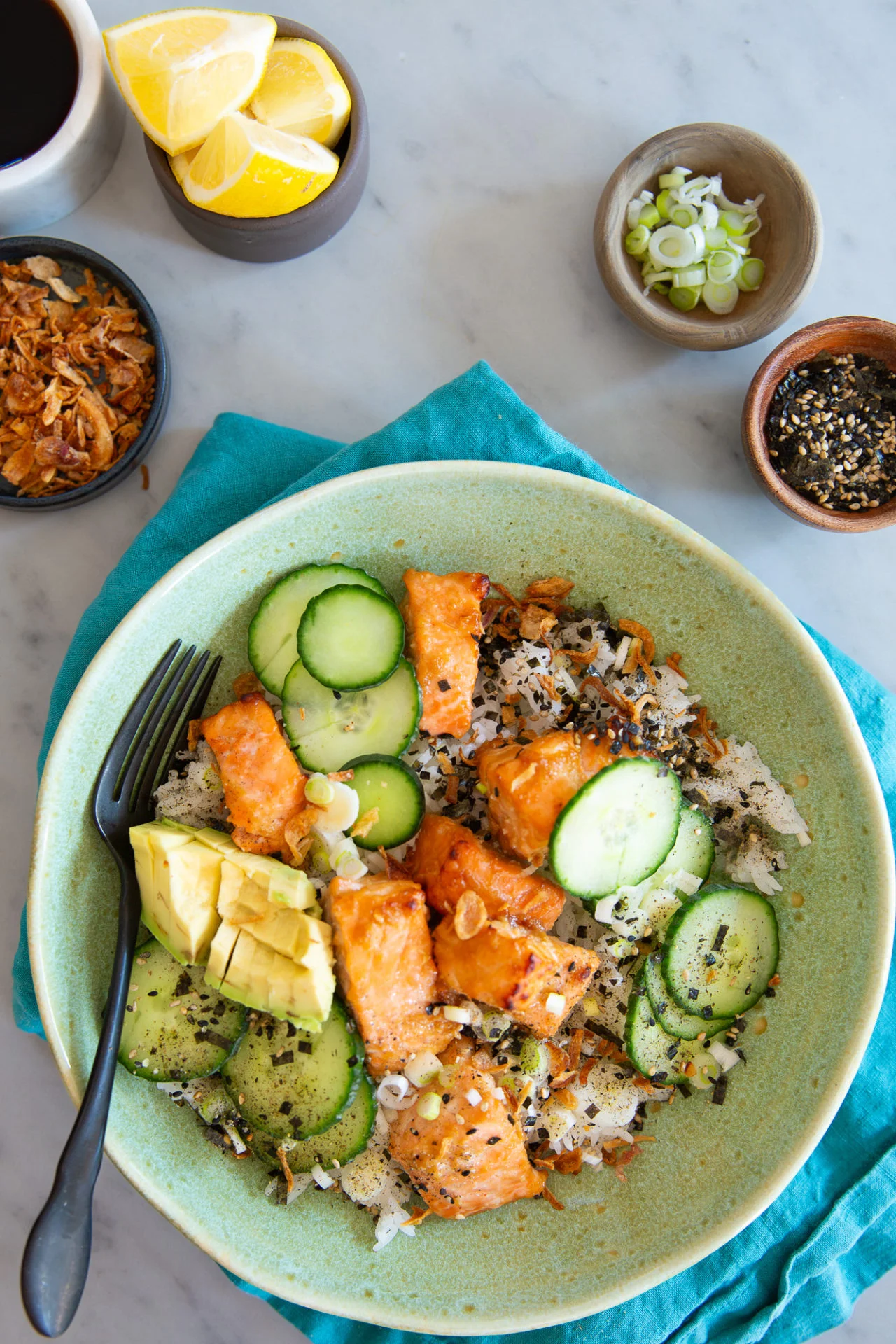 A salmon bowl featuring glazed miso salmon pieces, sliced cucumber, avocado, green onions, and sesame seeds. A black fork rests on the bowl. Around it sit dishes of lemon wedges, green onions, crispy onions, and sesame seeds.