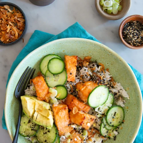 A Miso Salmon Bowl featuring rice, crispy miso salmon pieces, cucumber slices, avocado, green onions, and sesame seeds, garnished with fried onions. Surrounding the bowl are lemon wedges, extra green onions, fried onions, and sesame seeds.
