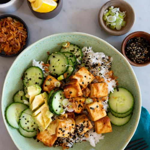 A green bowl holds a vibrant Tofu Bowl: rice topped with sautéed tofu cubes, sliced cucumber, avocado, scallions, black and white sesame seeds, and crispy shallots. Lemon wedges and soy sauce complete this fresh miso-inspired recipe on a marble surface.