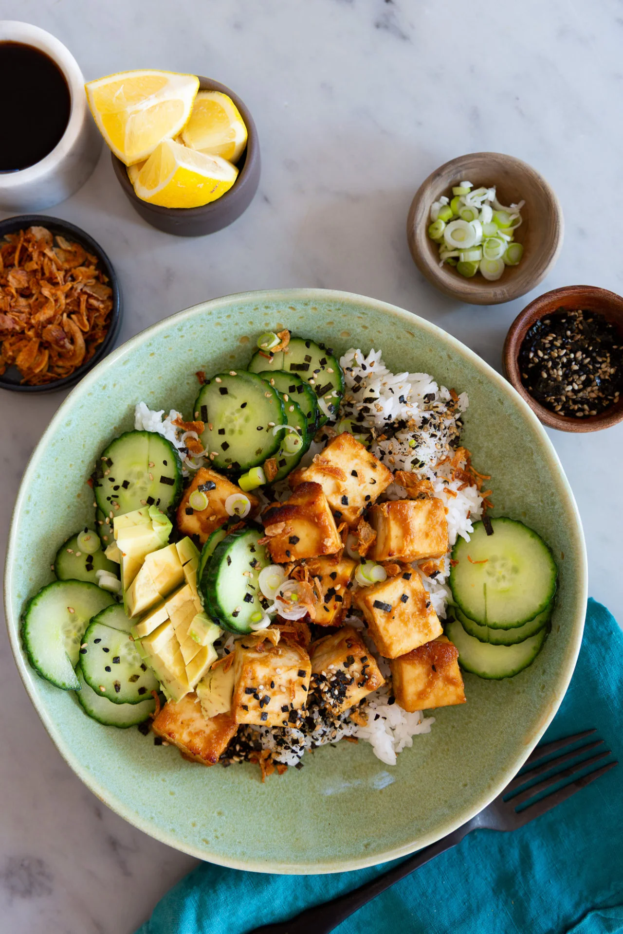 A green bowl holds a vibrant Tofu Bowl: rice topped with sautéed tofu cubes, sliced cucumber, avocado, scallions, black and white sesame seeds, and crispy shallots. Lemon wedges and soy sauce complete this fresh miso-inspired recipe on a marble surface.