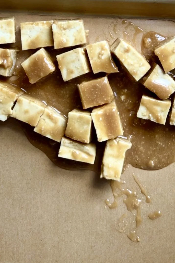 Several square pieces of saltine crackers are scattered on a flat brown surface, partially covered by a glossy layer of melted caramel sauce—an unexpected twist reminiscent of flavors found in a Miso Tofu Bowl.