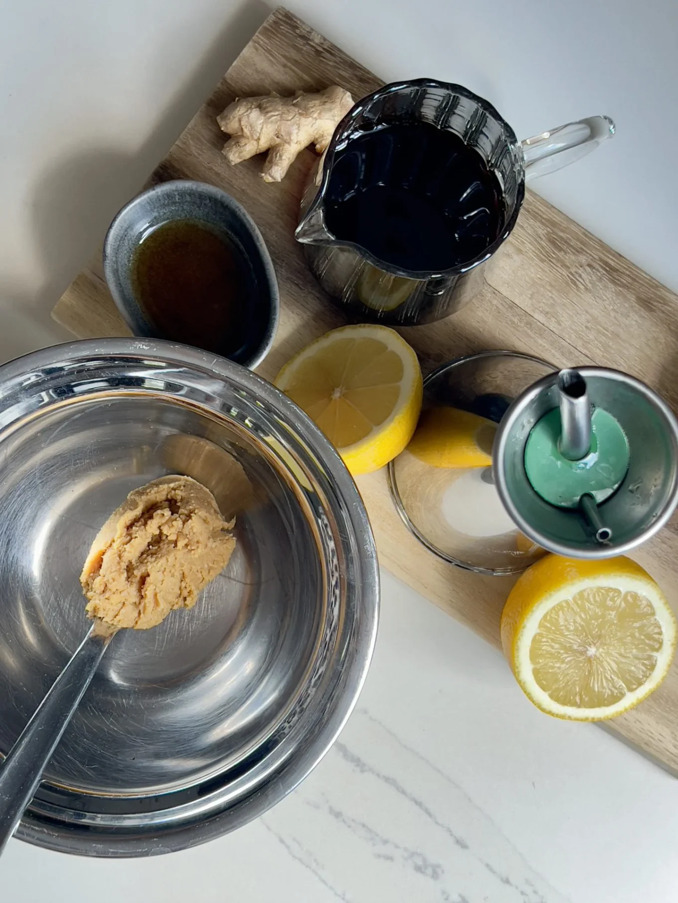 A metal bowl with a spoonful of miso paste sits on a counter beside halved lemons, soy sauce, fresh ginger, and oil—perfect ingredients for preparing a delicious Miso Salmon Bowl on a wooden cutting board.