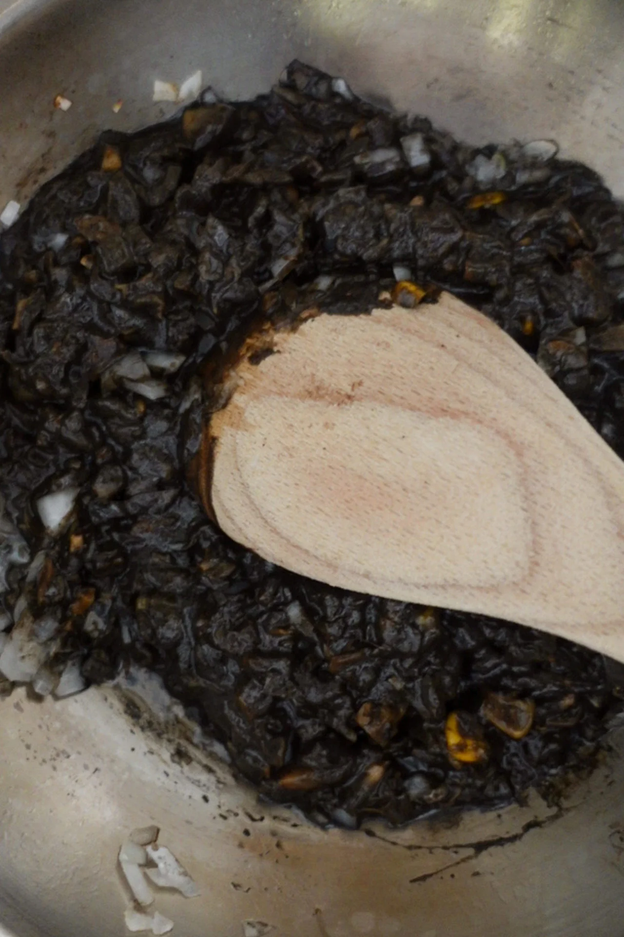 A close-up of a stainless steel pan in the kitchen holds a rich, dark mixture, likely Huitlacoche or Mexican truffle, with finely chopped vegetables and scattered white onion bits as a wooden spoon stirs the savory blend.