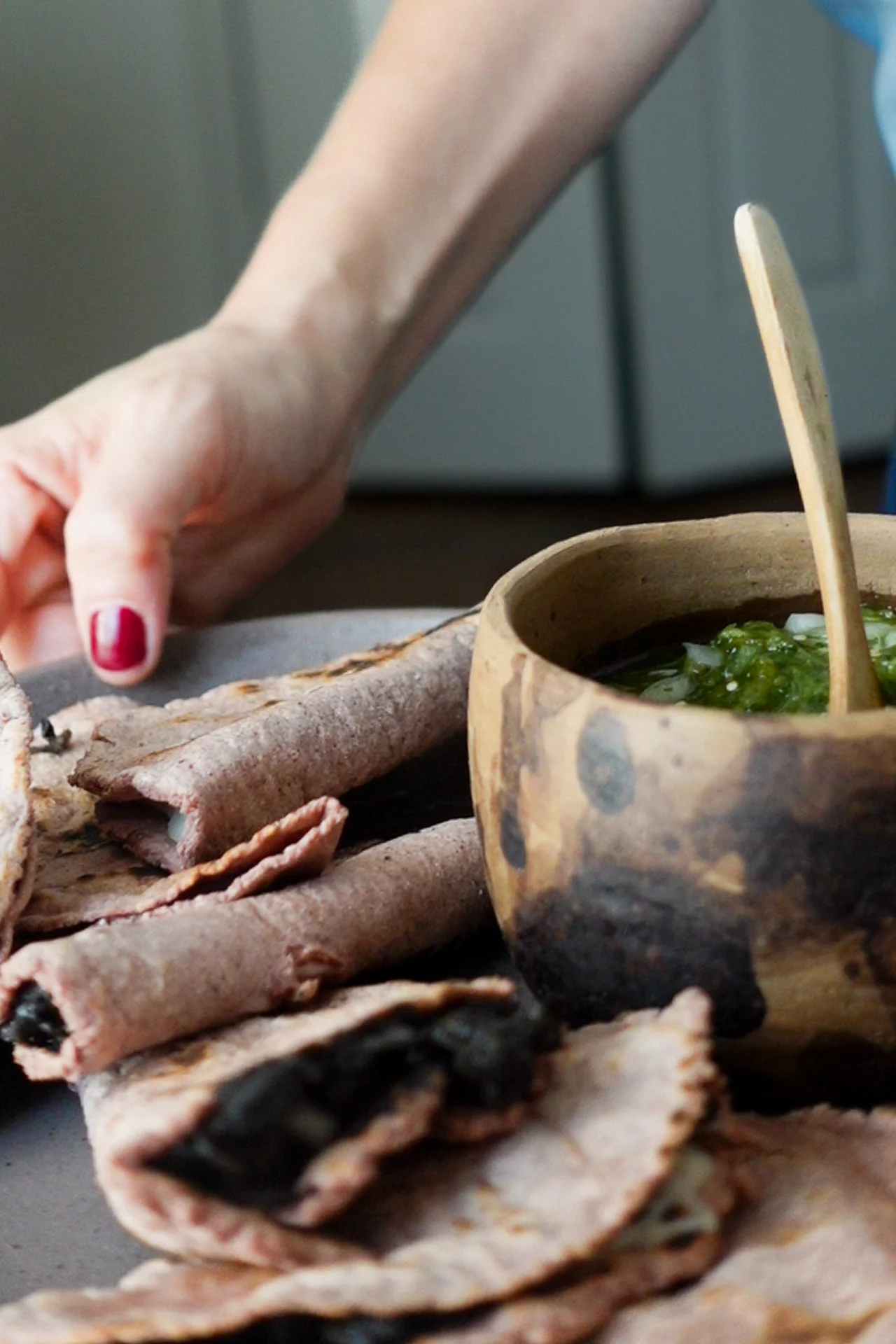 A hand with red nail polish reaches for a rolled, rustic blue corn tortilla—perfect for quesadillas—next to a wooden bowl filled with green salsa and a small wooden spoon. Several more tortillas are arranged around the bowl on a gray surface.