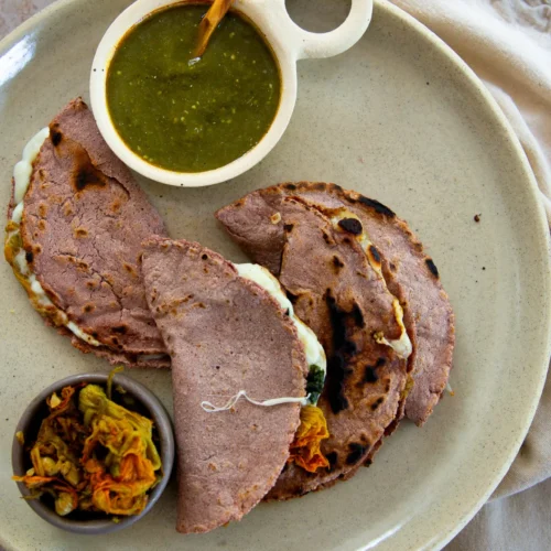 Three blue corn quesadillas with melted cheese are arranged on a beige plate. A small bowl of green salsa with a wooden spoon and another small bowl of pickled vegetables are also on the plate, which sits on a light fabric napkin on a stone surface.