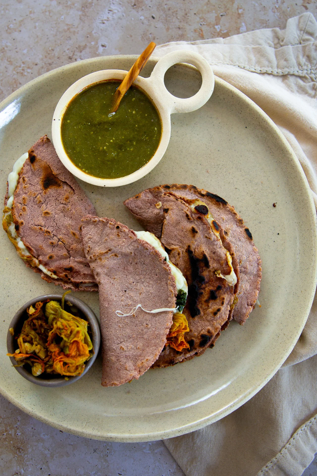 Three blue corn quesadillas with melted cheese are arranged on a beige plate. A small bowl of green salsa with a wooden spoon and another small bowl of pickled vegetables are also on the plate, which sits on a light fabric napkin on a stone surface.