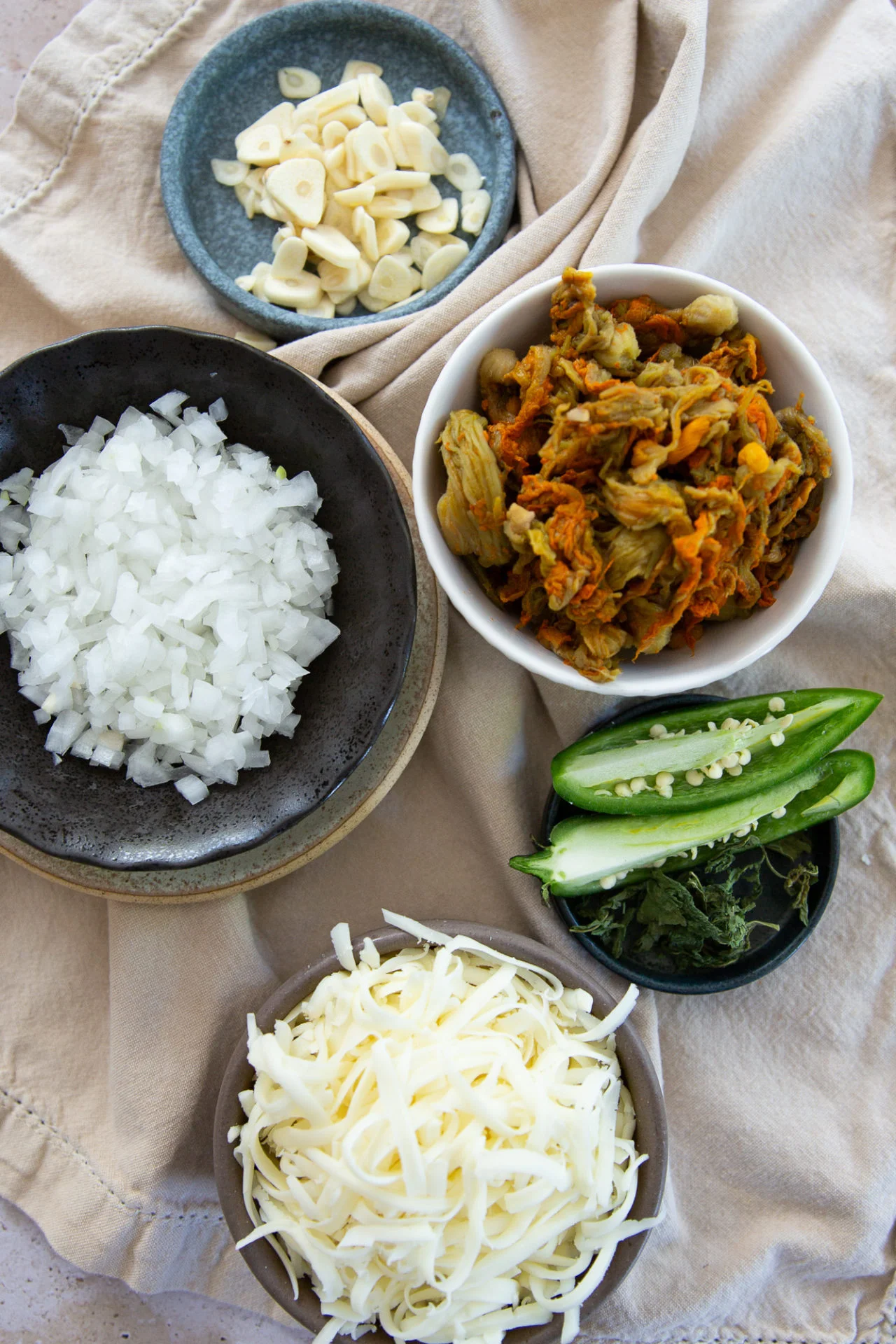 Five bowls on a beige cloth contain sliced garlic, chopped onions, shredded cheese, sliced jalapeños, and a kimchi mixture—ingredients perfectly prepped for Squash Blossom Quesadillas. Each is neatly arranged to showcase vibrant colors and textures from above.