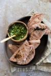 A dark oval plate holds five folded blue corn quesadillas, some with char marks, next to a small wooden bowl of chunky green salsa with chopped onions. The plate sits on a beige napkin atop a stone surface. Some quesadillas are filled with huitlacoche.