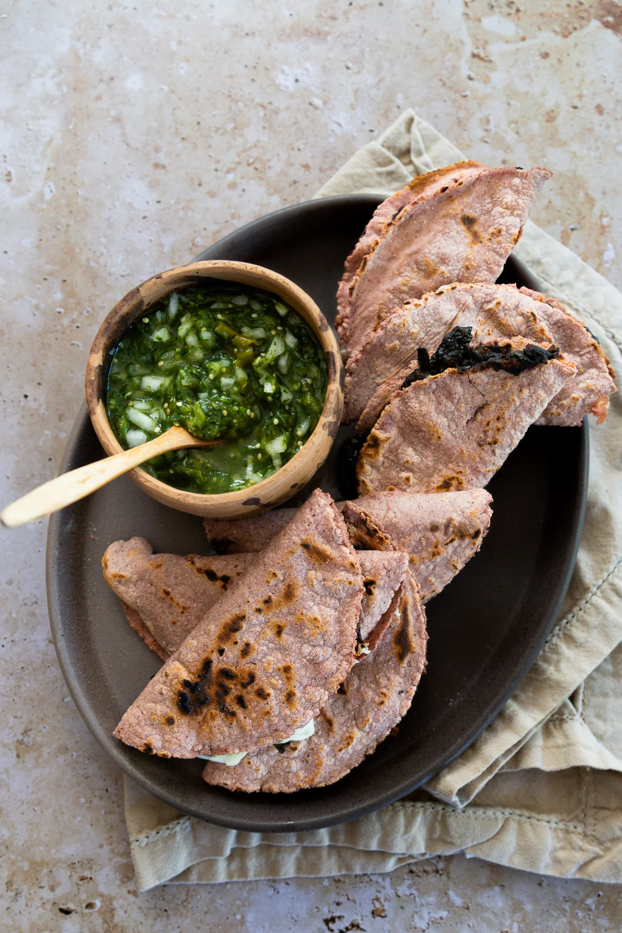 A dark oval plate holds five folded blue corn quesadillas, some with char marks, next to a small wooden bowl of chunky green salsa with chopped onions. The plate sits on a beige napkin atop a stone surface. Some quesadillas are filled with huitlacoche.