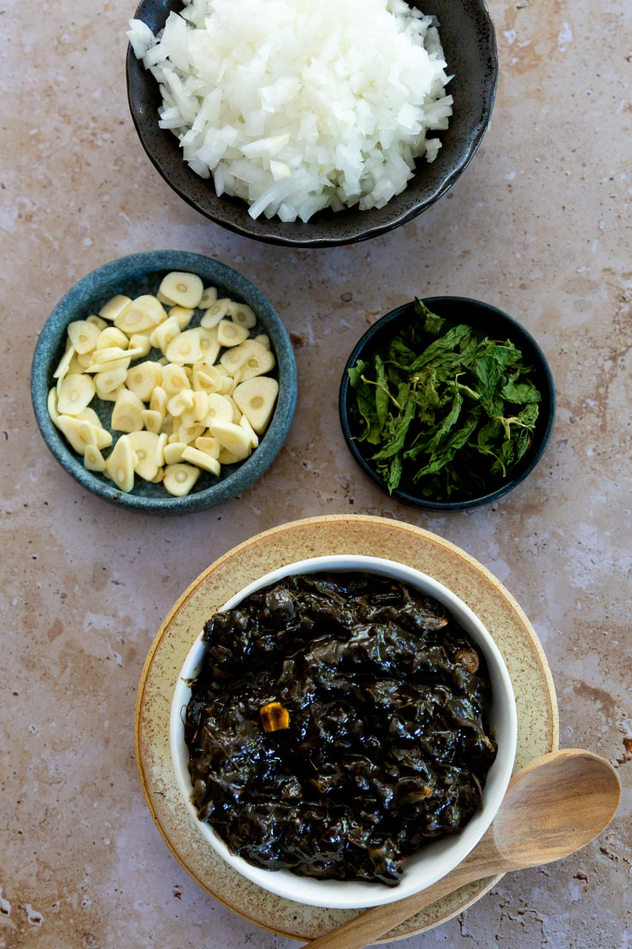 Four bowls on a tan stone surface: chopped white onions, sliced garlic, fresh herbs, and a saucy paste—classic ingredients for authentic Mexican recipes like quesadillas with huitlacoche. A wooden spoon rests beside the setup.