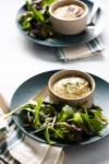 A blue plate holds a small ramekin of savory pie topped with browned mashed potatoes and herbs, alongside a fresh green salad. A gold fork rests beside the food. In the background, a second plate and ramekin with salad are partially visible.