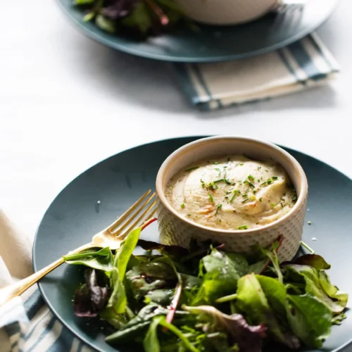 A blue plate holds a small ramekin of savory pie topped with browned mashed potatoes and herbs, alongside a fresh green salad. A gold fork rests beside the food. In the background, a second plate and ramekin with salad are partially visible.