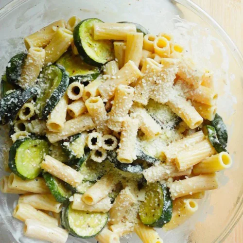 A glass bowl of fresh zucchini pasta salad with rigatoni, sliced cooked zucchini, and a sprinkle of parmesan sits on a white countertop next to a wooden cutting board, all lightly coated in a creamy parmesan dressing.