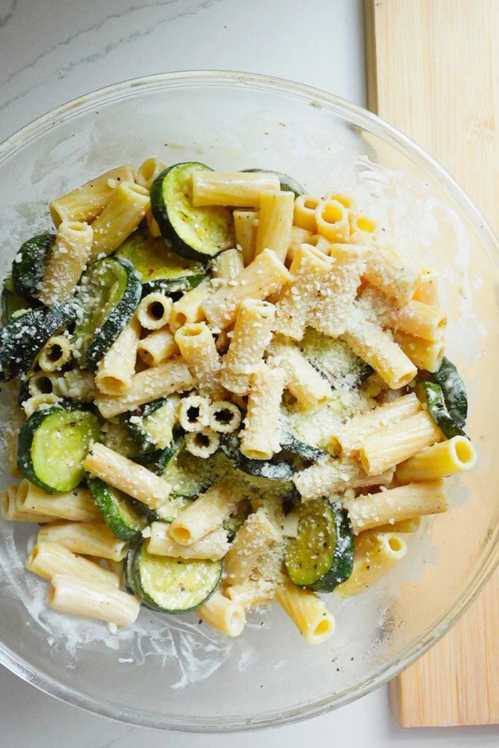 A glass bowl of fresh zucchini pasta salad with rigatoni, sliced cooked zucchini, and a sprinkle of parmesan sits on a white countertop next to a wooden cutting board, all lightly coated in a creamy parmesan dressing.