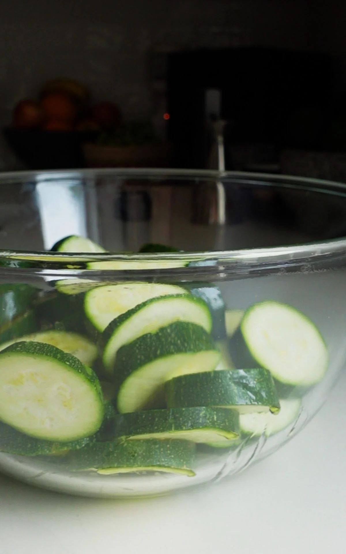 A glass bowl filled with thickly sliced green zucchini pieces sits on a white countertop, perfect for a Fresh Zucchini Pasta Salad. The vibrant slices are ready to be tossed with a Parmesan Dressing. A blurred bowl of fruit and kitchen items can be seen behind.